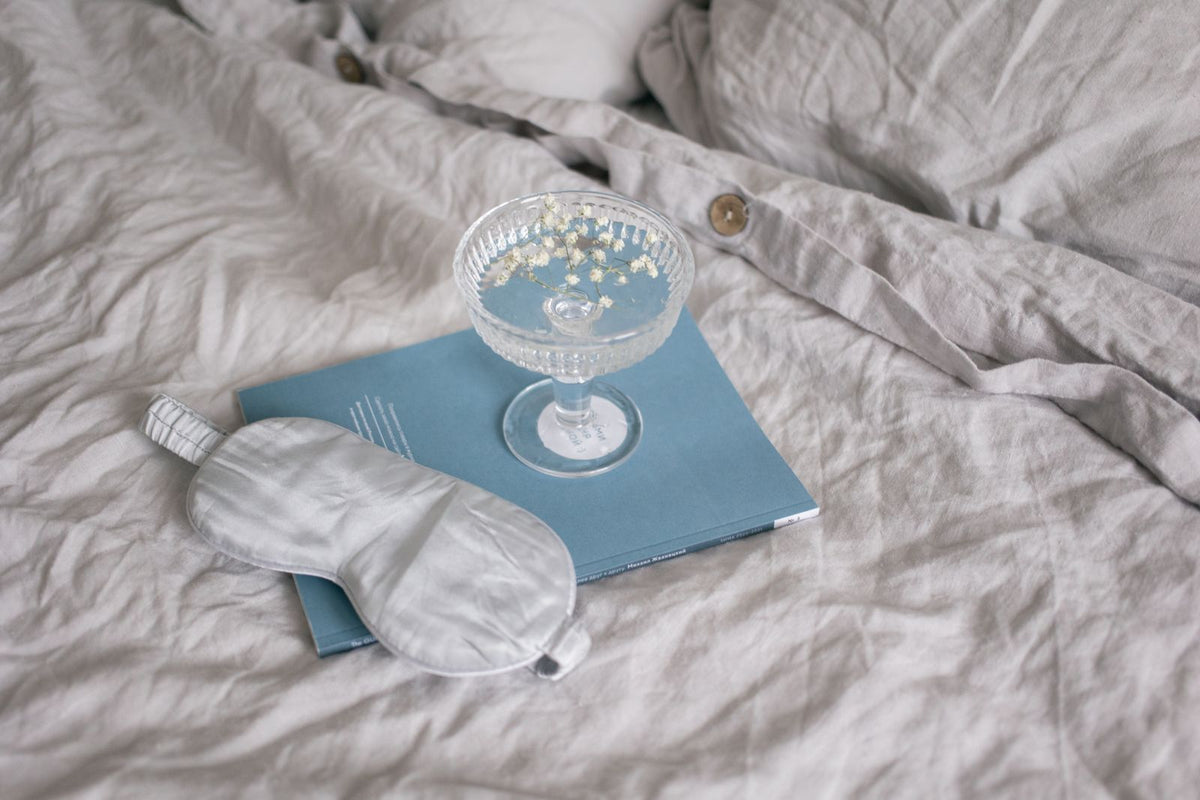 A sleep mask and glass of white flowers on a blue book, set on cozy bedding