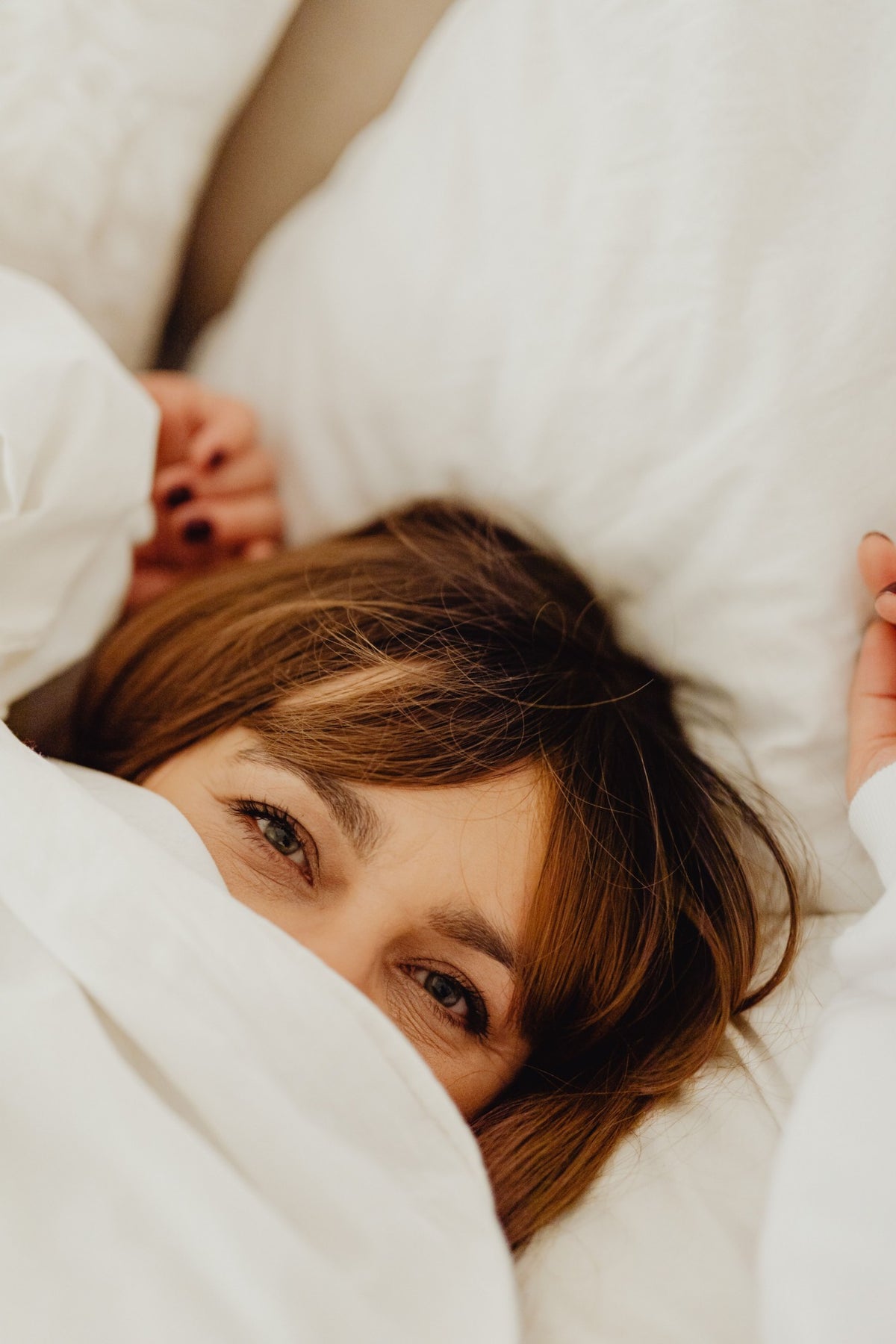 A woman peeking out from under white bedding, her eyes smiling at the camera